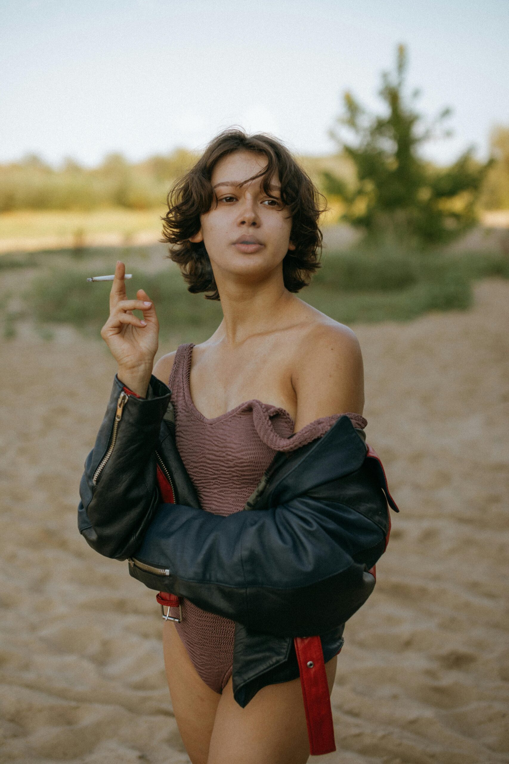 Young woman in a swimsuit with a leather jacket, holding a cigarette on a sandy beach.